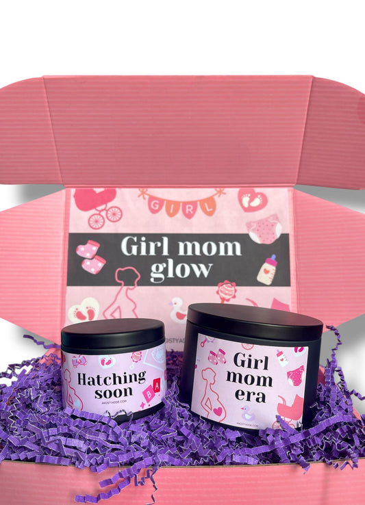 A pink cardboard box with a pink and white design, containing two candles labeled "Hatching soon" and "Girl mom era".