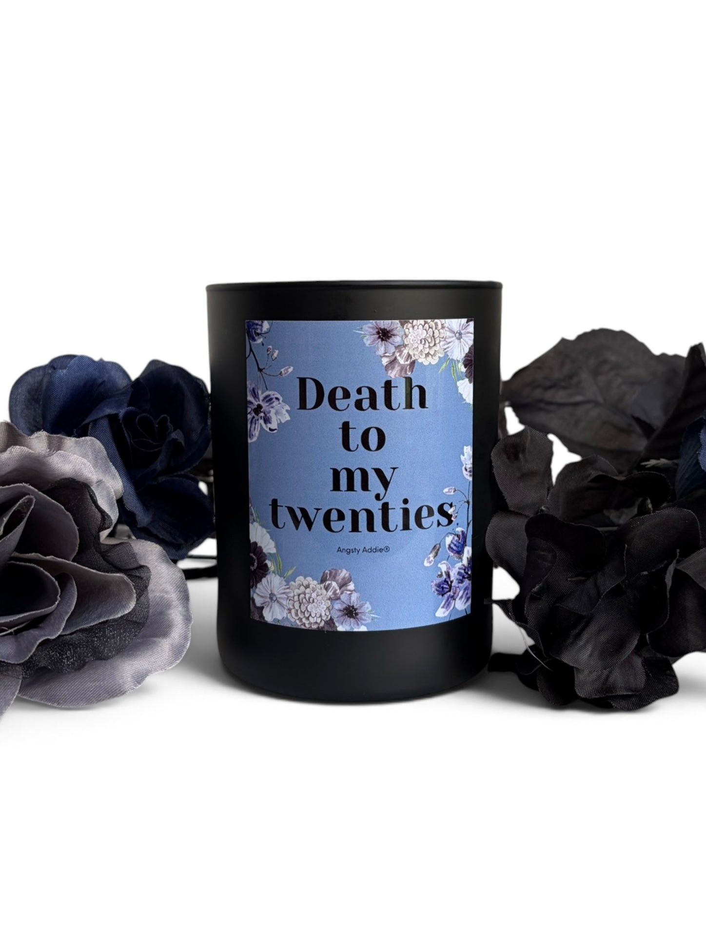 A black glass candle jar with a light blue floral label reading 'Death to my twenties' by Angela Abbott, styled with dark dried flowers on a white background.